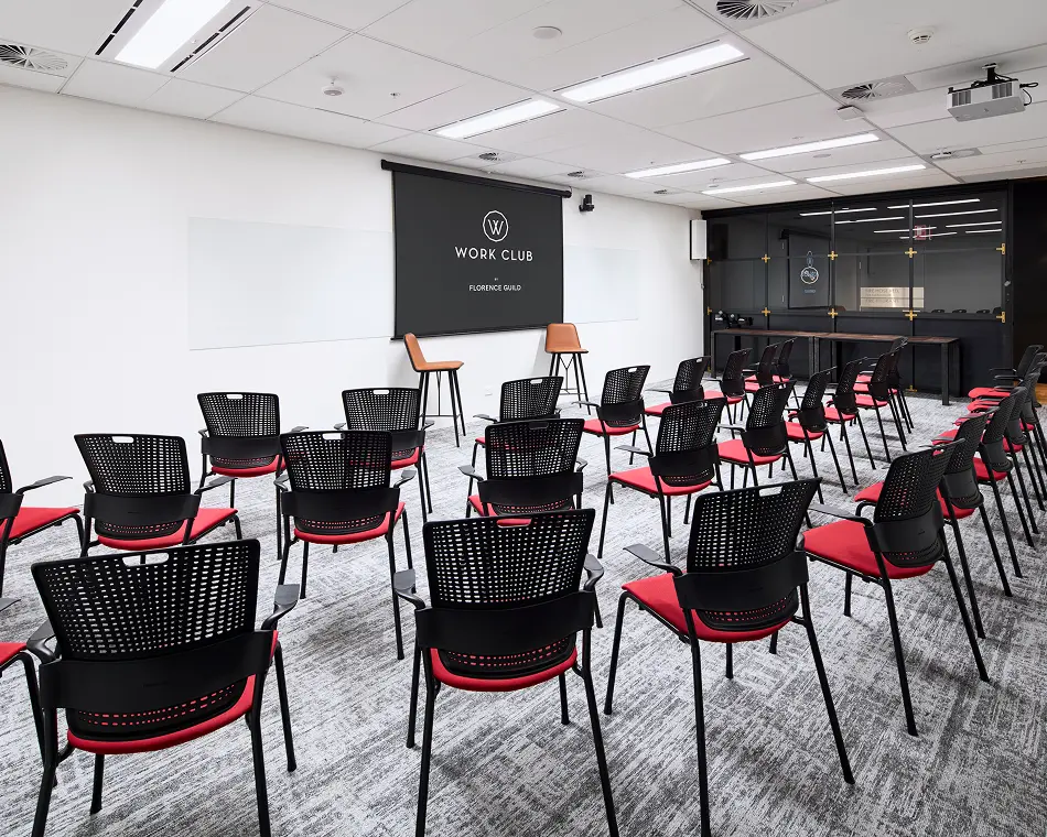 Multiple rows of black and red chairs face a central presentation screen inside a bright, modern conference room with carpeted floors and white walls. The text on the screen reads: "W" "WORK CLUB" "BY" "FLORENCE GUILD"