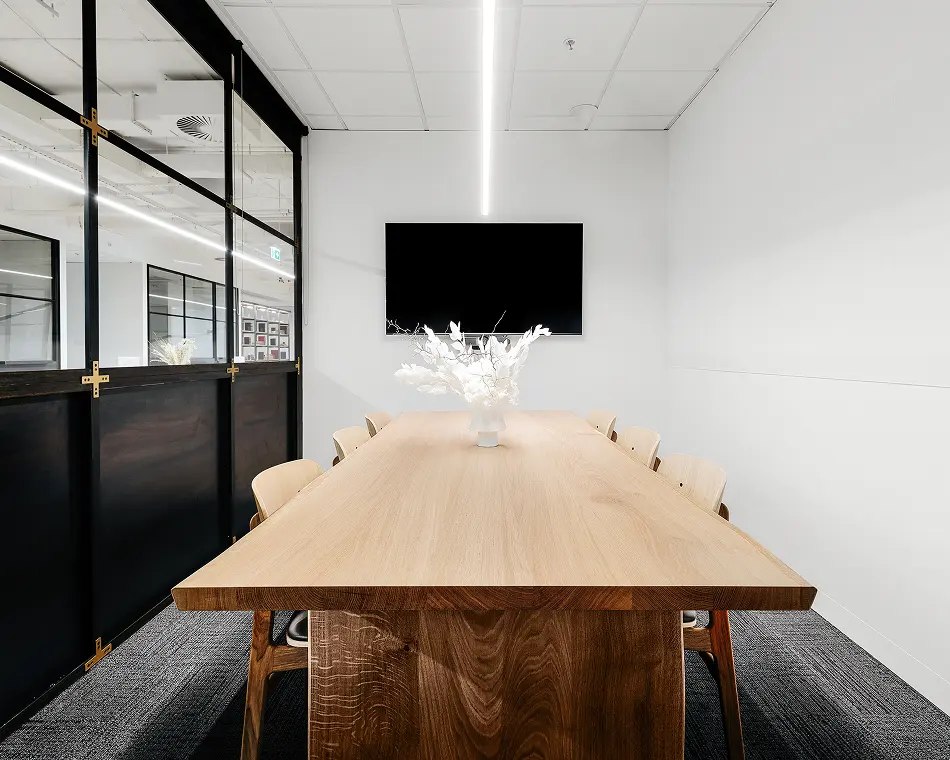 A long wooden conference table lined with chairs sits beneath a linear light, featuring a white centerpiece, inside a modern meeting room with white walls and a glass partition.