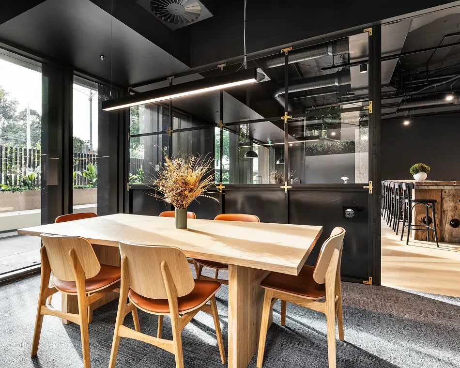 A light wooden table surrounded by brown leather-seated chairs stands under a suspended linear light inside a modern office featuring dark glass partitions and large floor-to-ceiling windows.