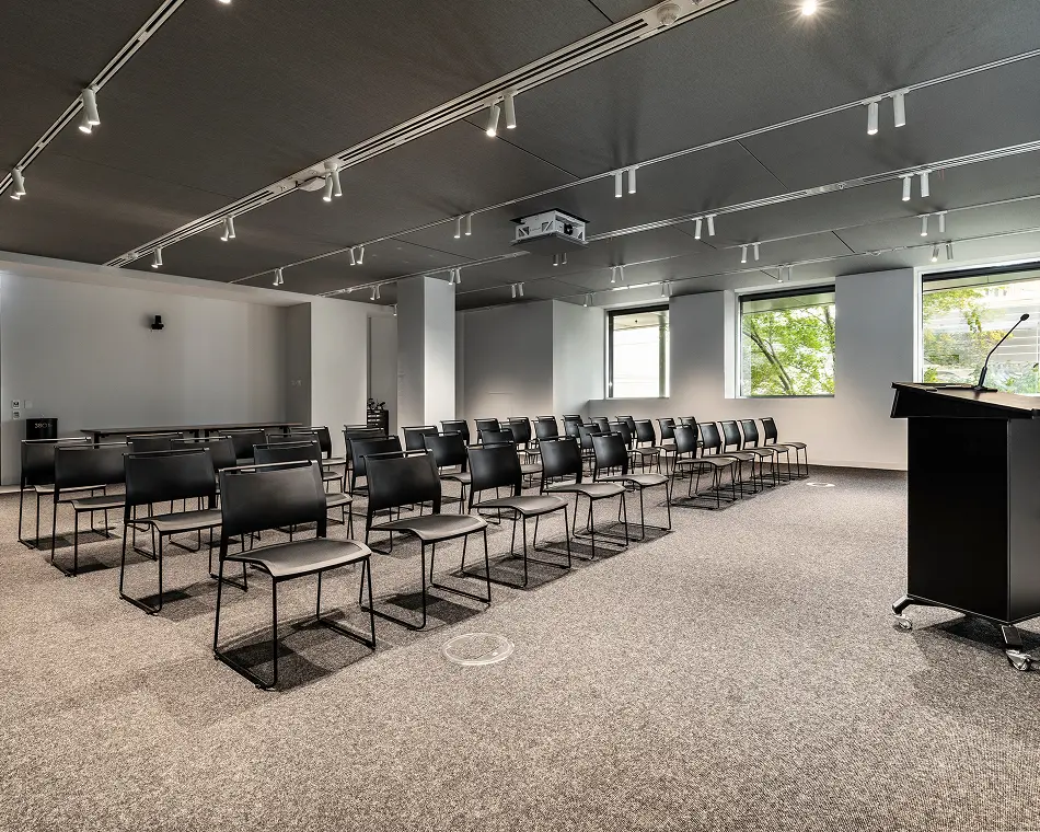 Neat rows of black chairs face a black podium in a modern presentation room with grey carpet, track lighting on a dark ceiling, and windows overlooking greenery.