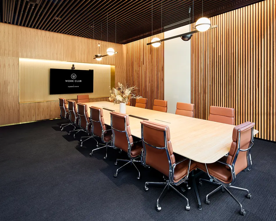 A long light-wood conference table surrounded by cognac leather chairs sits in a modern boardroom with vertical wood-slat walls and a black-slatted ceiling. A wall-mounted screen displays a "W WORK CLUB" logo, beside a dried floral arrangement beneath pendant lights.