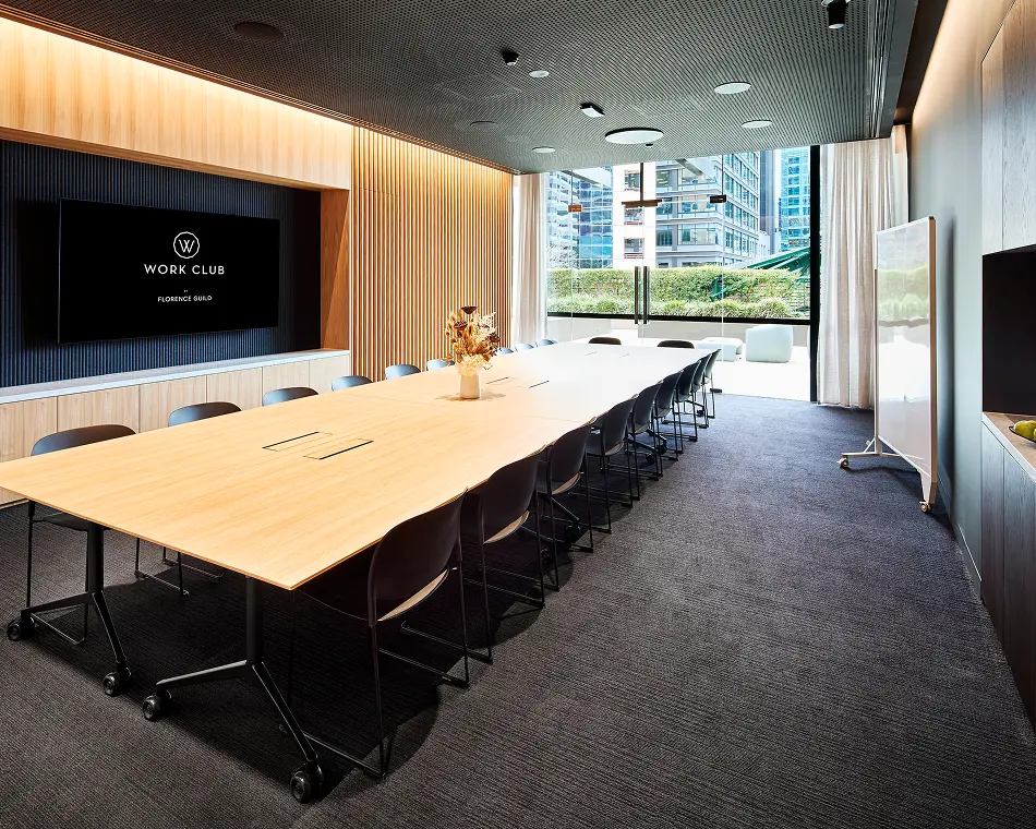 A long wooden conference table surrounded by black chairs fills a modern boardroom featuring wood-paneled walls, dark carpet, a whiteboard, and glass doors opening to a terrace. The screen displays: "W WORK CLUB BY FLORENCE GUILD".