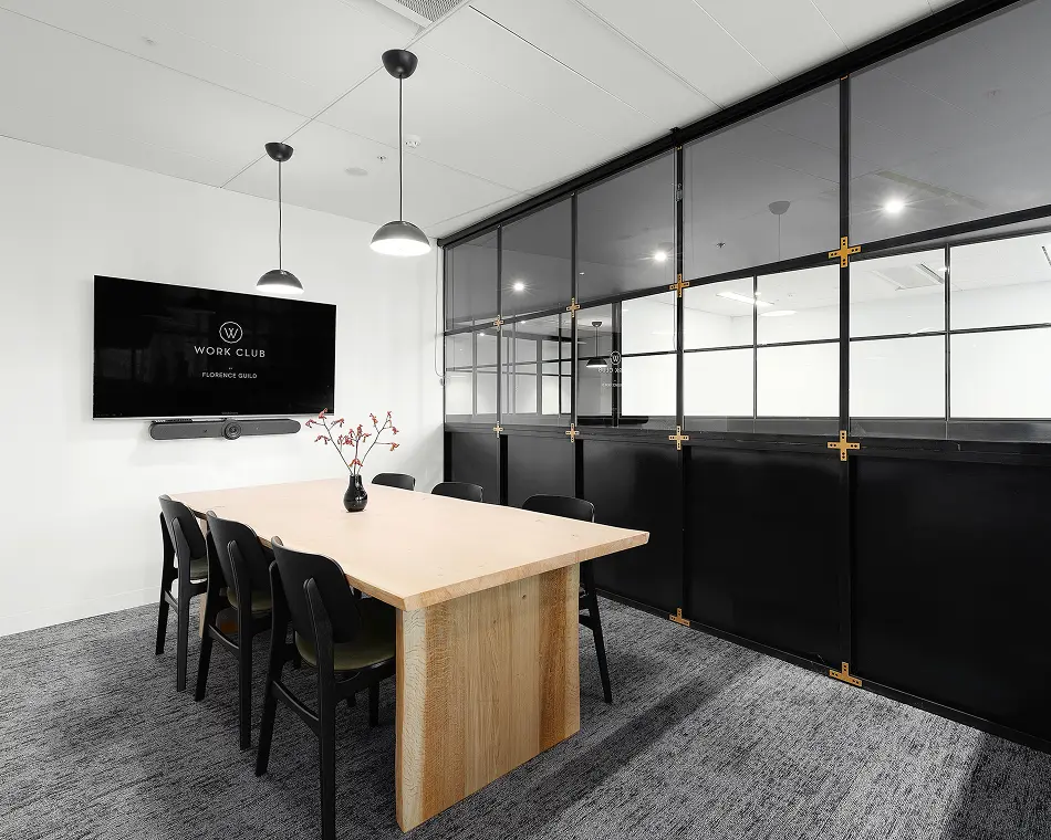 A light-wood conference table surrounded by black chairs sits under pendant lights in a modern meeting room, facing a wall-mounted monitor displaying "W WORK CLUB BY FLORENCE GUILD".