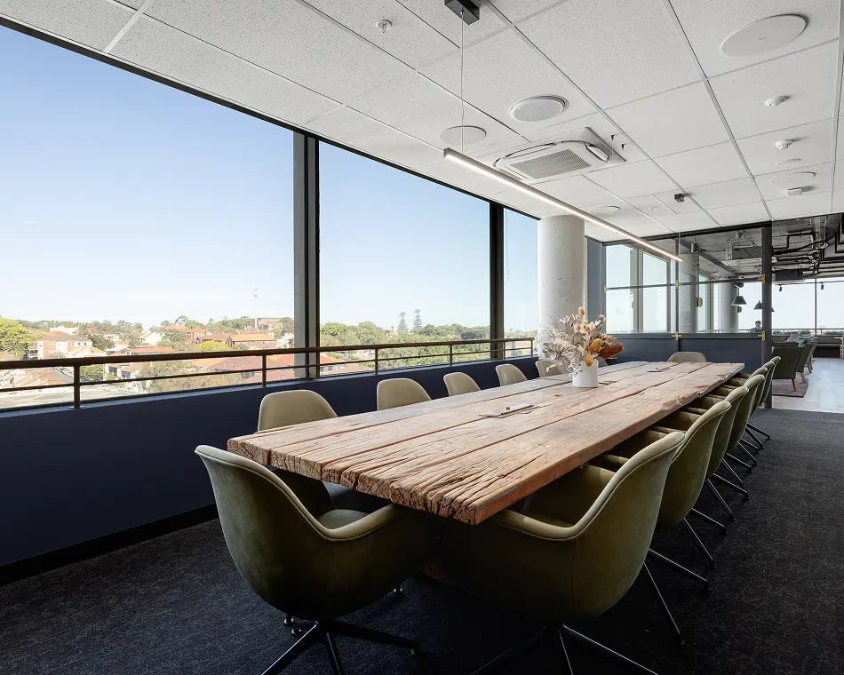 A long, rustic wooden conference table surrounded by olive-green swivel chairs sits inside a modern boardroom, positioned next to floor-to-ceiling windows overlooking a sunny suburban landscape.