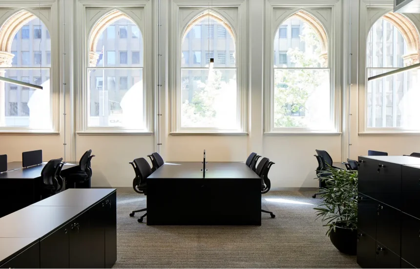 Black desks and office chairs are arranged in a modern carpeted office facing five large arched windows that look out onto city buildings and trees.