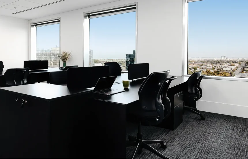 Sleek black desks paired with ergonomic chairs sit inside a modern office room featuring large windows that overlook a sprawling cityscape under a bright, clear sky.