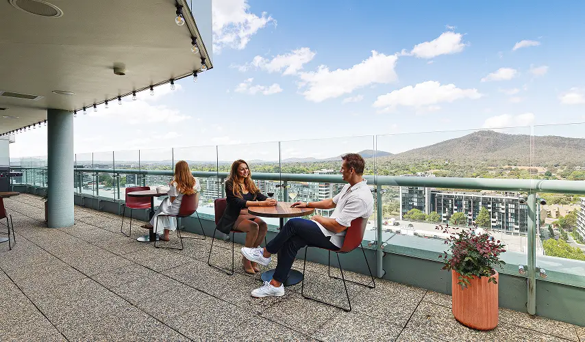 A man and a woman sit conversing at a small table on a high-rise balcony, overlooking an expansive city landscape and distant hills under a bright, partly cloudy sky.