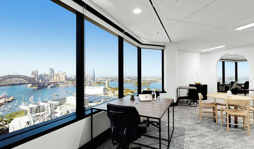 Wooden office desks and chairs sit beside expansive floor-to-ceiling windows, offering a panoramic view of a sunny harbor city, iconic bridge, and skyline.
