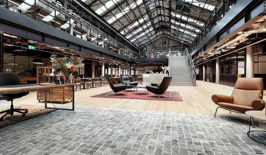 A spacious, modern industrial office atrium features leather armchairs, wooden desks, and a wide central staircase beneath a high vaulted glass-and-steel ceiling, with a "W" logo on the reception desk.