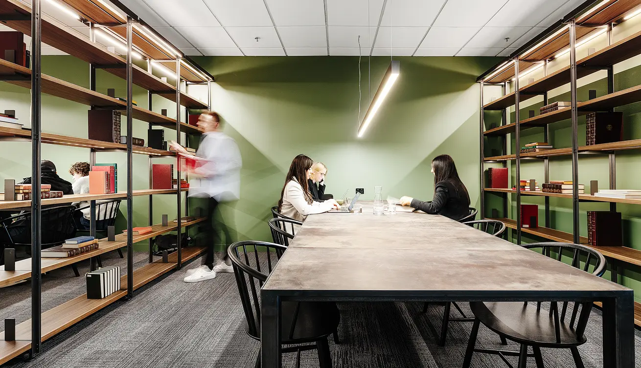 People work at a long wooden table while a blurred man walks past tall, illuminated bookshelves lining a modern study room featuring olive-green walls and dark carpet.