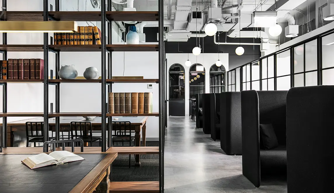 Open wooden bookshelves lined with books separate study desks from high-backed black seating booths along a polished concrete hallway in a modern office with exposed ceiling pipes and globe pendant lights.