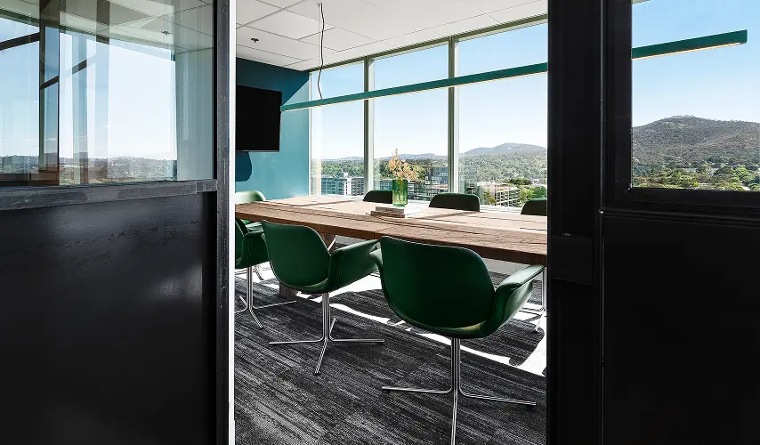 Green swivel chairs surround a long wooden conference table inside a modern office room featuring floor-to-ceiling windows that overlook a city landscape and mountains.