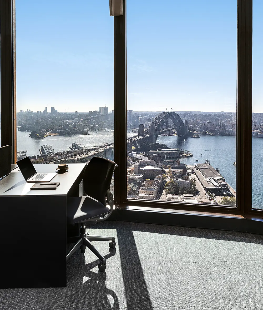 An office desk with an open laptop and chair sits beside floor-to-ceiling windows, offering a panoramic high-rise view of the Sydney Harbour Bridge and surrounding cityscape.