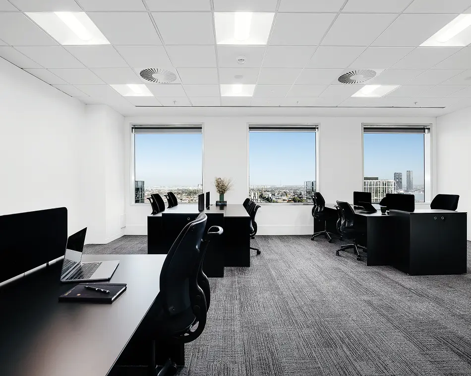Modern black office desks and ergonomic chairs are neatly arranged inside a bright, white-walled office featuring grey carpeted flooring and large windows overlooking a sprawling city view.