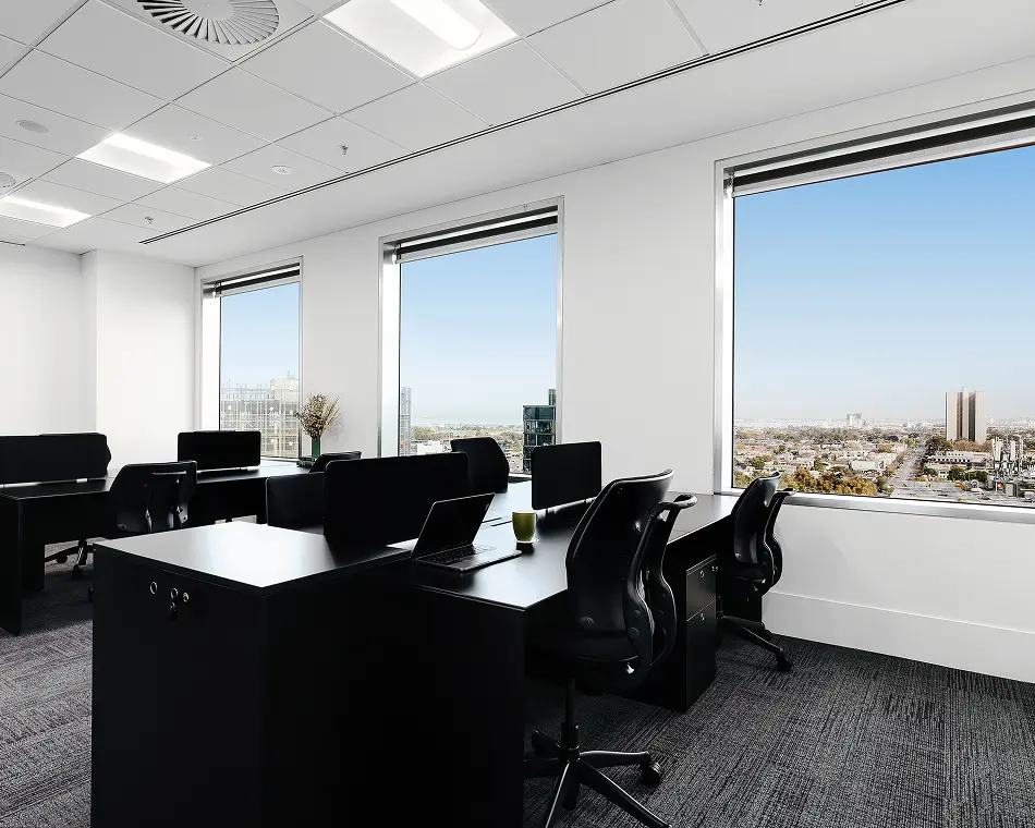 Black office workstations with ergonomic chairs sit arranged inside a bright room beside large windows that reveal an expansive view of a city skyline under clear skies.
