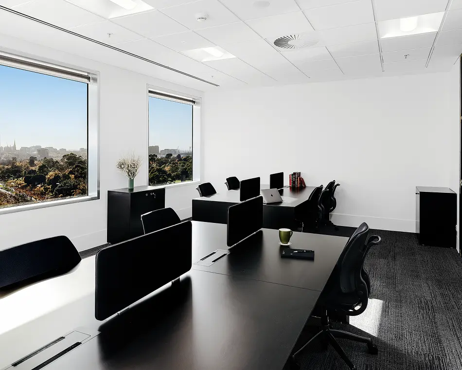 Black modern office desks paired with ergonomic chairs sit arranged in a spacious room featuring white walls, dark carpeting, and large windows looking out onto a park and cityscape.