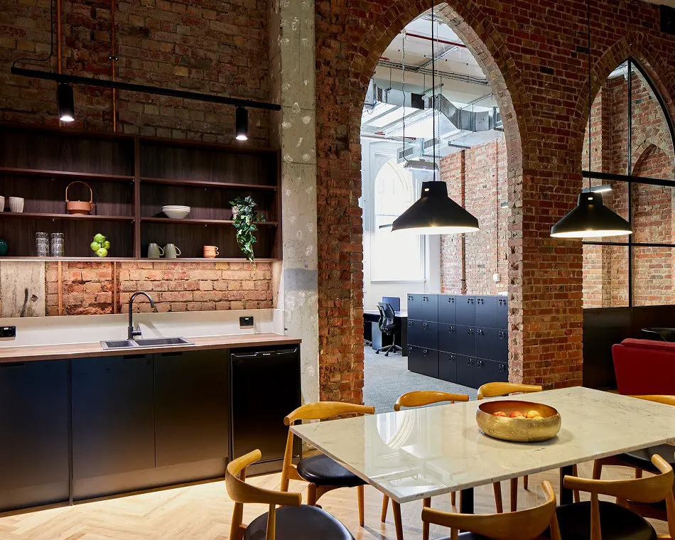 A marble dining table surrounded by wooden chairs stands in a modern kitchen, featuring dark cabinetry under open shelving, set against exposed brick walls with archways leading to office spaces.