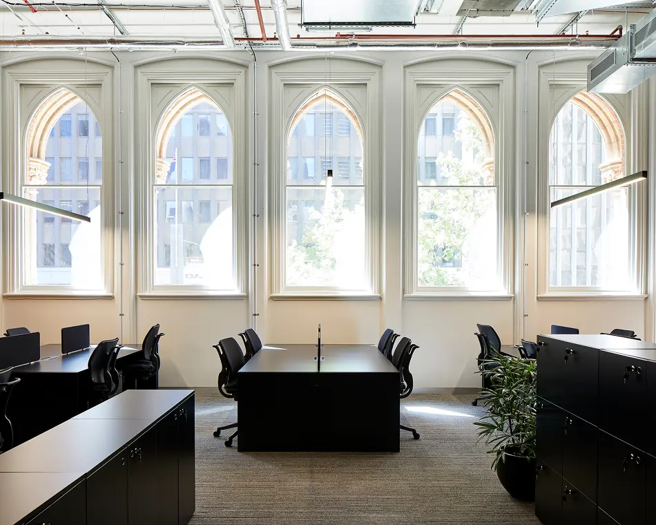 Black office desks and rolling chairs sit neatly arranged inside a modern office space, framed by five large arched windows looking out onto neighboring city buildings.