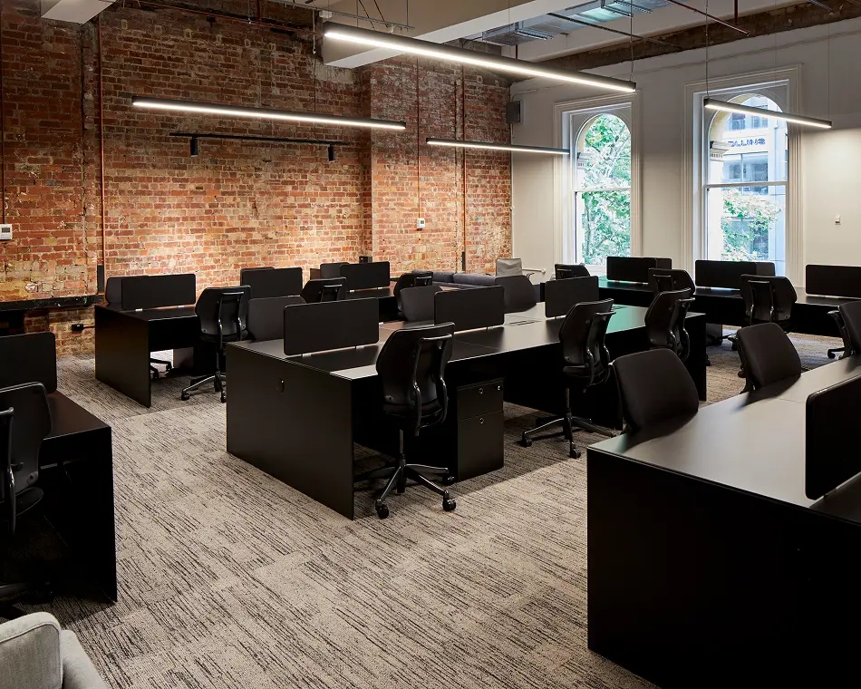 Sleek black desks paired with office chairs sit arranged in a modern open-plan workspace featuring carpeted floors, hanging linear lights, exposed red-brick walls, and arched windows.