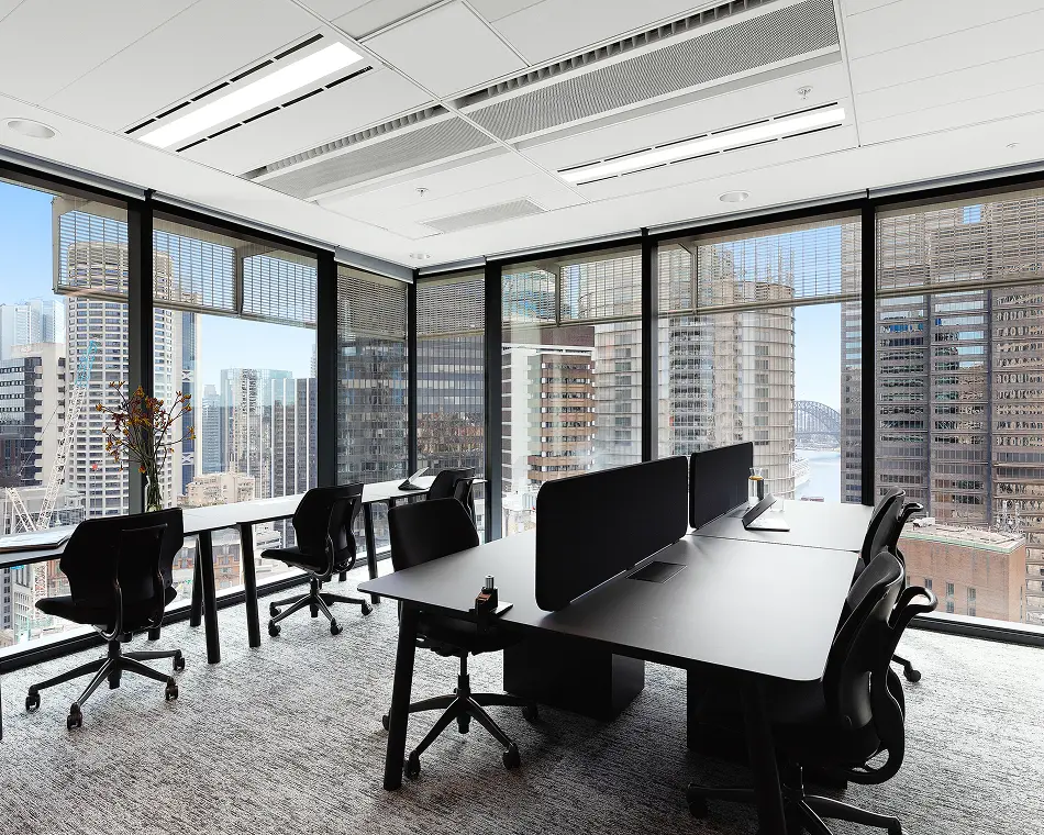Black office desks with ergonomic chairs sit inside a carpeted workspace, framed by floor-to-ceiling glass windows overlooking a panoramic city skyline and skyscrapers.