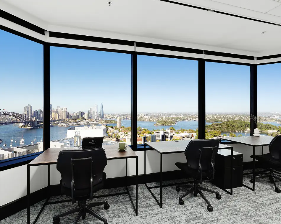 Black office chairs and wooden desks face floor-to-ceiling windows overlooking a panoramic harbor cityscape. A laptop on one desk displays text that reads: WORK CLUB.