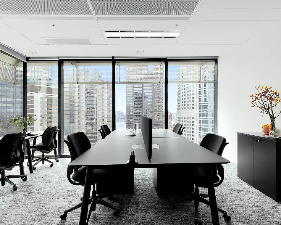 A long dark conference desk flanked by black office chairs sits in a modern high-rise office, featuring expansive floor-to-ceiling windows that offer views of a towering city skyline.