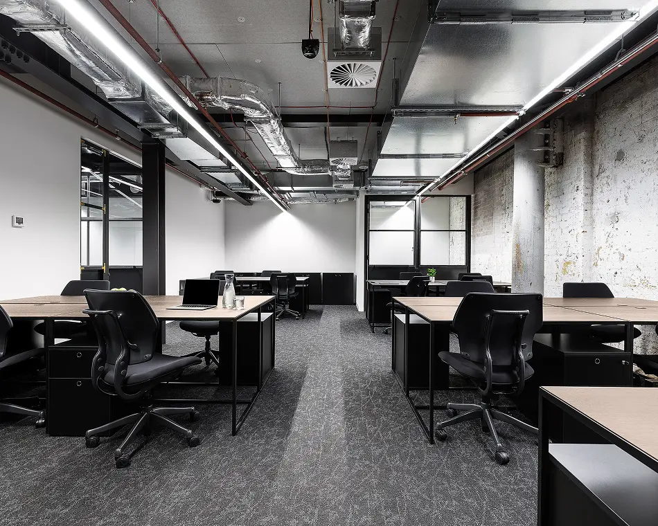 Black ergonomic chairs and wooden desks stand arranged in rows inside a modern industrial office space, featuring dark carpet, exposed overhead metal ductwork, linear lights, and a concrete wall.