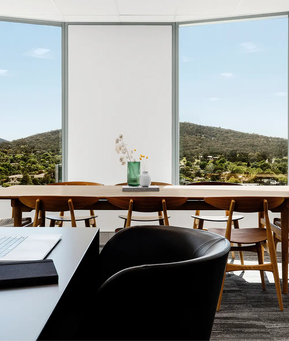 A long wooden conference table with chairs sits before floor-to-ceiling windows overlooking forested hills, while a black desk occupies the foreground. Text on the book reads "CURATE".