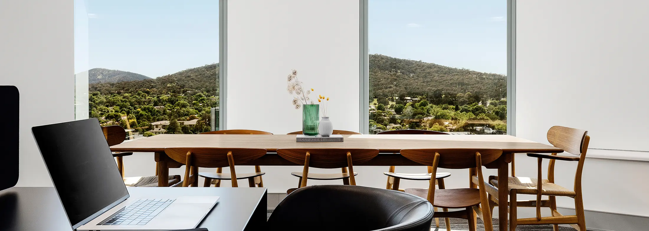 A long wooden table surrounded by chairs stands inside a modern room featuring large windows that reveal a scenic, tree-covered mountain landscape. The text "CURATE" and "INSPIRATION FOR AN INDIVIDUAL HOME" is visible on the book spine resting on the table.