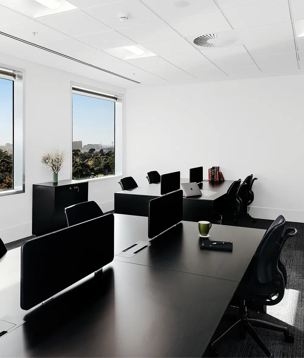 Modern black office desks with privacy screens and ergonomic chairs stand neatly arranged inside a bright, minimalist white office featuring large windows overlooking a city skyline.