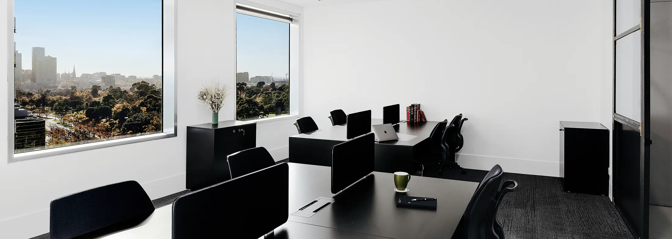Black office workstations fitted with monitors and ergonomic chairs sit arranged inside a bright, white-walled office featuring large windows overlooking a distant city skyline and park.