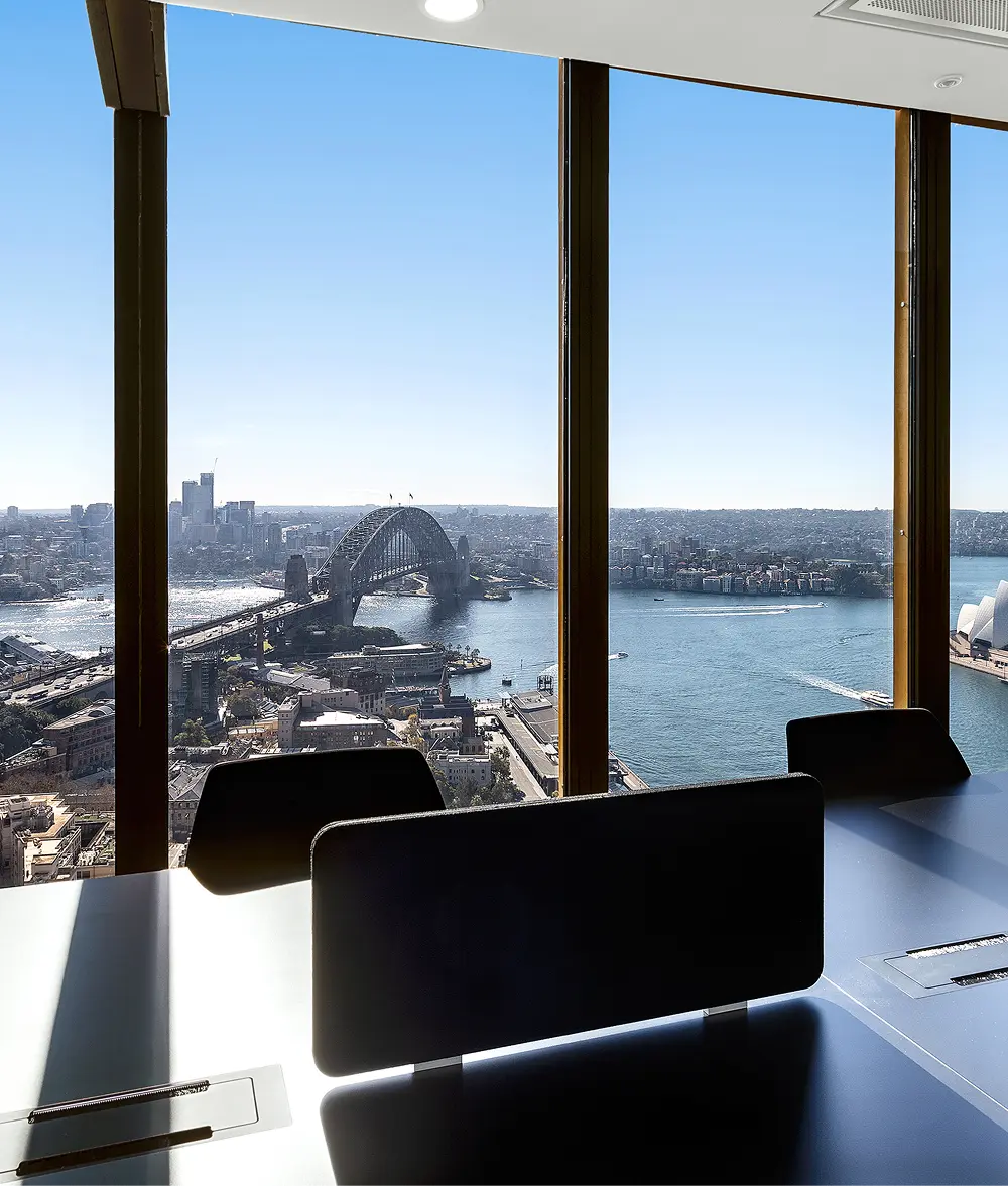 Black office chairs sit around a conference table before floor-to-ceiling windows framing a high-rise panoramic view of the Sydney Harbour Bridge and sparkling water under a clear blue sky.