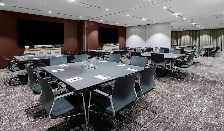 Square gray conference tables surrounded by dark chairs stand neatly arranged with water pitchers and note pads inside a spacious, carpeted meeting room featuring wall-mounted monitors.