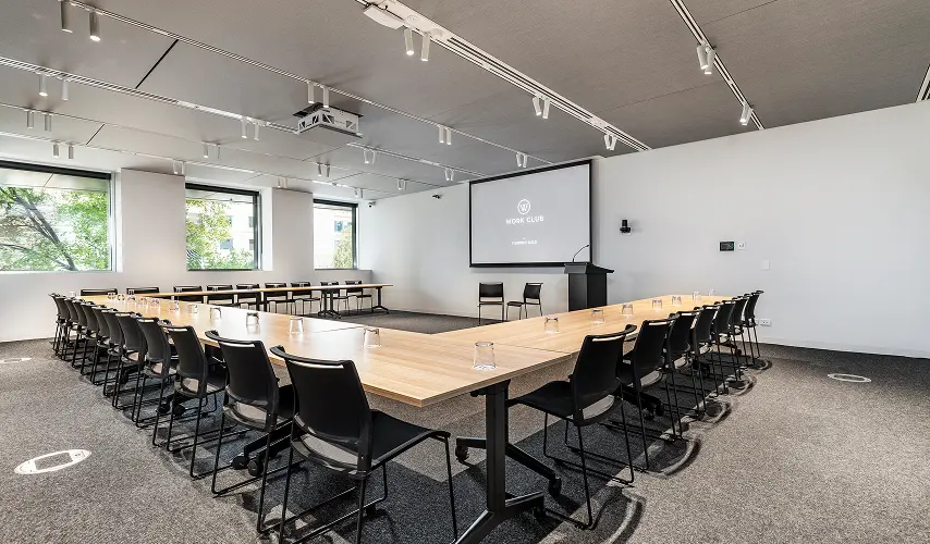 A large U-shaped wooden table setup with black chairs fills a modern conference room facing a presentation screen and podium. Text on the screen reads: “W WORK CLUB BY FLORENCE GUILD”.