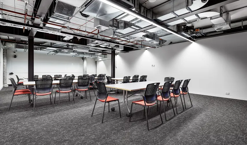 Black mesh chairs with orange seats surround long white tables, arranged in a modern conference room featuring exposed industrial ceiling ductwork, linear lights, and dark grey carpeted flooring.