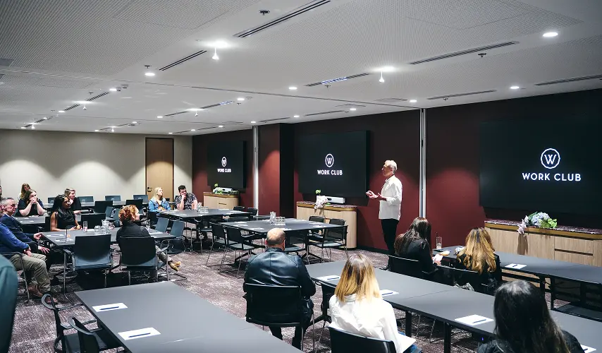 A speaker presents to an audience seated at black tables inside a conference room with dark red walls and large screens displaying a logo with the text "WORK CLUB".