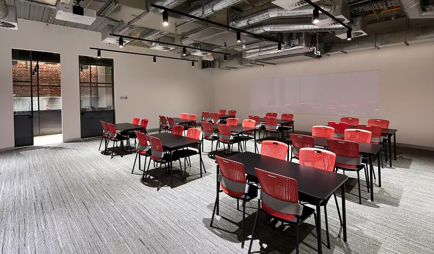 Black rectangular tables paired with bright red chairs are arranged across a carpeted room featuring exposed ceiling ductwork, track lighting, and a large whiteboard on the wall.