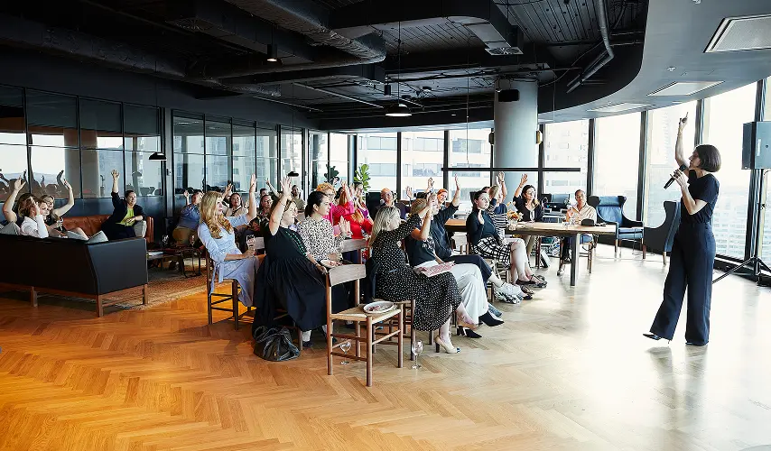 A woman holding a microphone addresses a seated audience raising their hands inside a modern, high-rise office room featuring floor-to-ceiling windows and herringbone wooden floors.